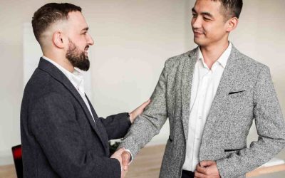 Un hombre occidental trajeado con barba y un hombre asiático también trajeado se dan la mano tras cerrar un trato para la importación de productos superando los aranceles y otras barreras al comercio internacional.